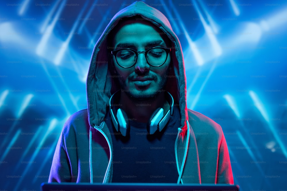 Junger Hacker in Kapuzenpullover und Brille bricht in Computersystem vor blau beleuchtetem Hintergrund mit geometrischen Linien ein