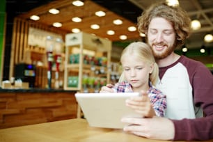 Young man and his daughter addicted to modern gadgets searching for curious videos or talking through video-chat at leisure