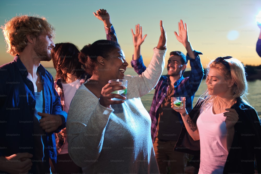 Cheerful girls with mojito and guys dancing together by seaside