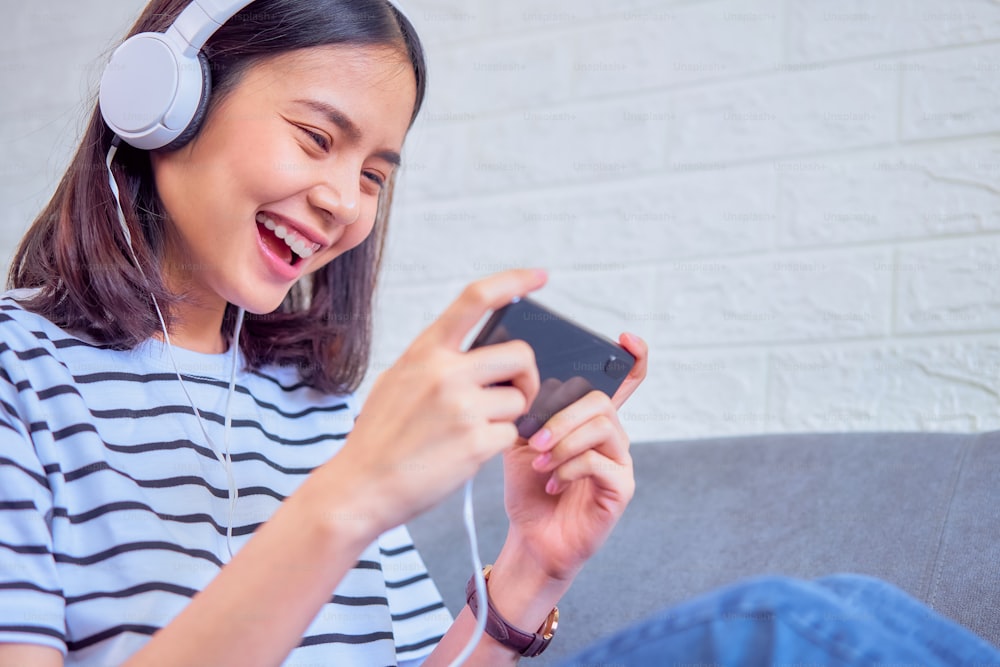 Aufgeregte junge asiatische Frau, die auf dem Sofa sitzt, trägt weiße Kopfhörer auf dem Kopf und spielt Spiele auf dem Smartphone im Wohnzimmer zu Hause.