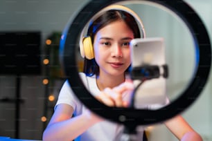 Smiling Young Asian woman wearing headset and live broadcasting on internet and reading comments with people in social media on smartphone.