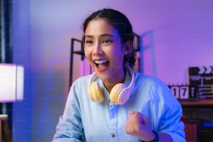 Emocionada joven asiática con auriculares y jugando en línea con los puños cerrados celebrando la victoria expresando éxito.