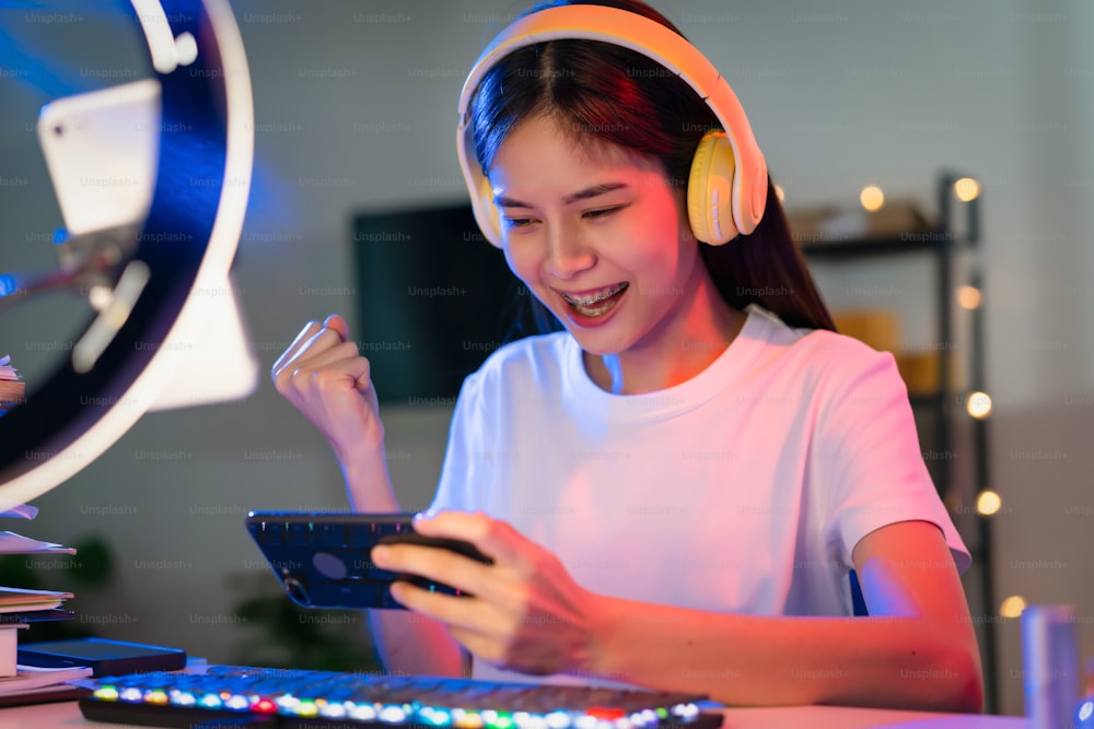 Excitada jovem asiática mulher usando fone de ouvido e jogando jogo on-line no smartphone com transmissão ao vivo na internet.