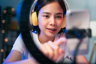 Sonriente joven asiática con auriculares y transmisión en vivo en Internet y leyendo comentarios con personas en las redes sociales en un teléfono inteligente.