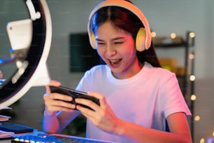 흥분한 젊은 아시아 여성들은 헤드셋을 쓰고 스마트폰으로 온라인 게임을 하며 인터넷 생방송을 한다.