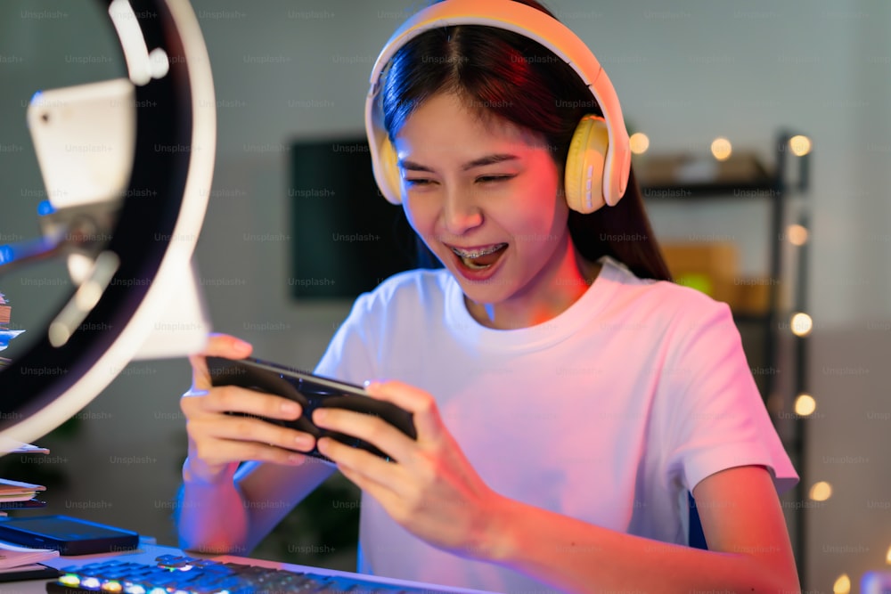 Aufgeregte junge asiatische Frau, die Headset trägt und Online-Spiel auf dem Smartphone mit Live-Übertragung im Internet spielt.