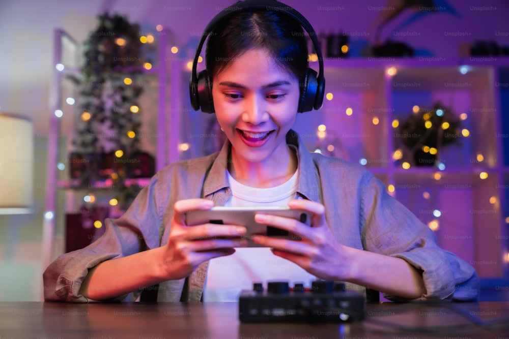 Emocionada joven asiática con auriculares y jugando en línea en un teléfono inteligente con transmisión en vivo por Internet.