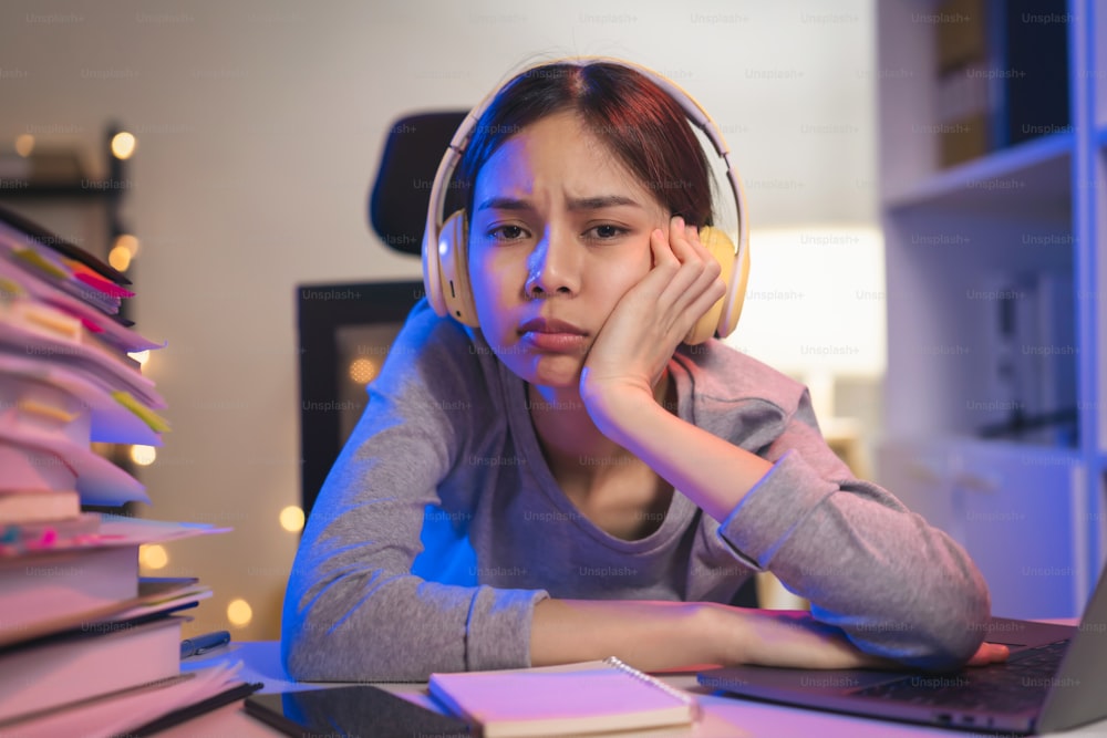 Stressige junge asiatische Frau, die ein Headset trägt und mit einem Stapel Papiere auf dem Tisch auf dem Stuhl sitzt und nachts auf den Computerbildschirm schaut.