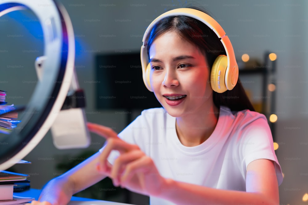 Jovem asiática sorridente usando fone de ouvido e transmissão ao vivo na internet e lendo comentários com pessoas nas mídias sociais no smartphone.