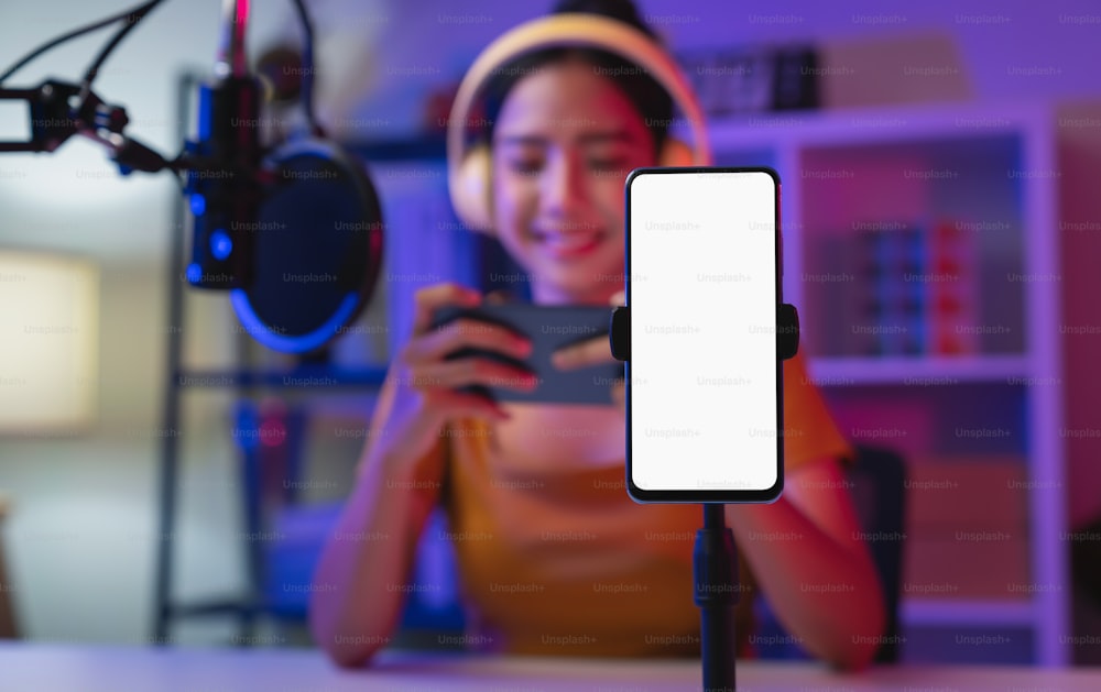 Cheerful beautiful Asian woman wearing headphones with live streaming on mobile and smartphone mockup of blank screen for application.