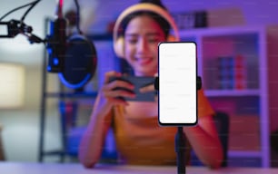 Cheerful beautiful Asian woman wearing headphones with live streaming on mobile and smartphone mockup of blank screen for application.