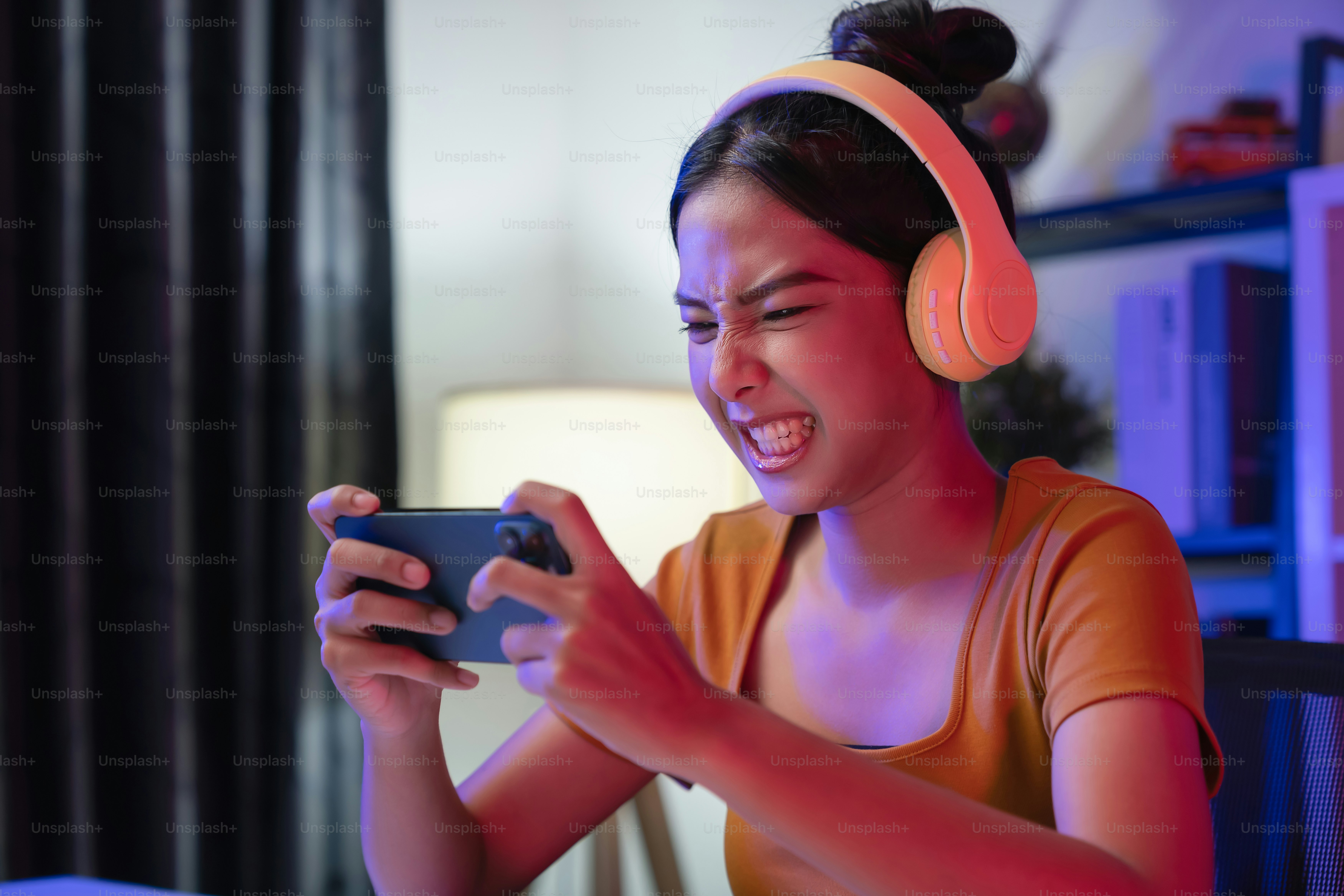 Wanita Asia muda yang bersemangat mengenakan headset dan bermain game online di smartphone di malam hari rumah modern.