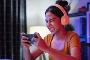 Excited Young Asian woman wearing headset and playing online game on smartphone at night modern house.