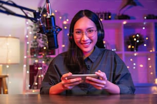 Emocionada joven asiática con auriculares y jugando en línea en un teléfono inteligente con transmisión en vivo por Internet.