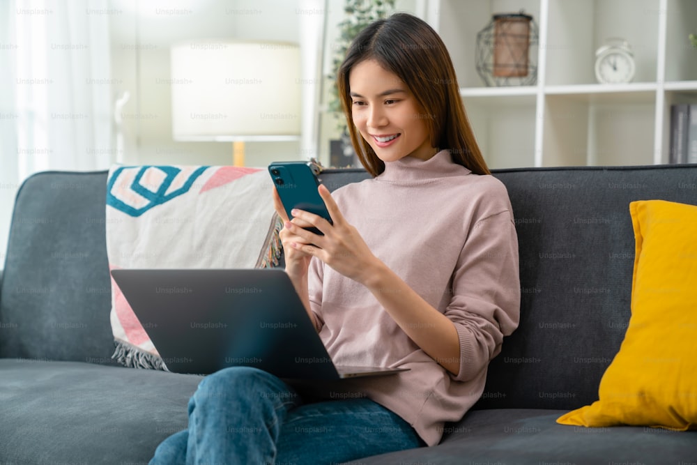 Young cheerful woman sitting on sofa and holding smartphone with using laptop computer at home.