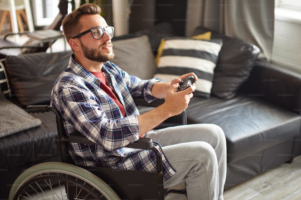 Giovane disabile che gioca ai videogiochi, seduto su sedia a rotelle a casa. Indossa occhiali e camicia a quadri. Qurantine, restate a casa, concetto di riabilitazione
