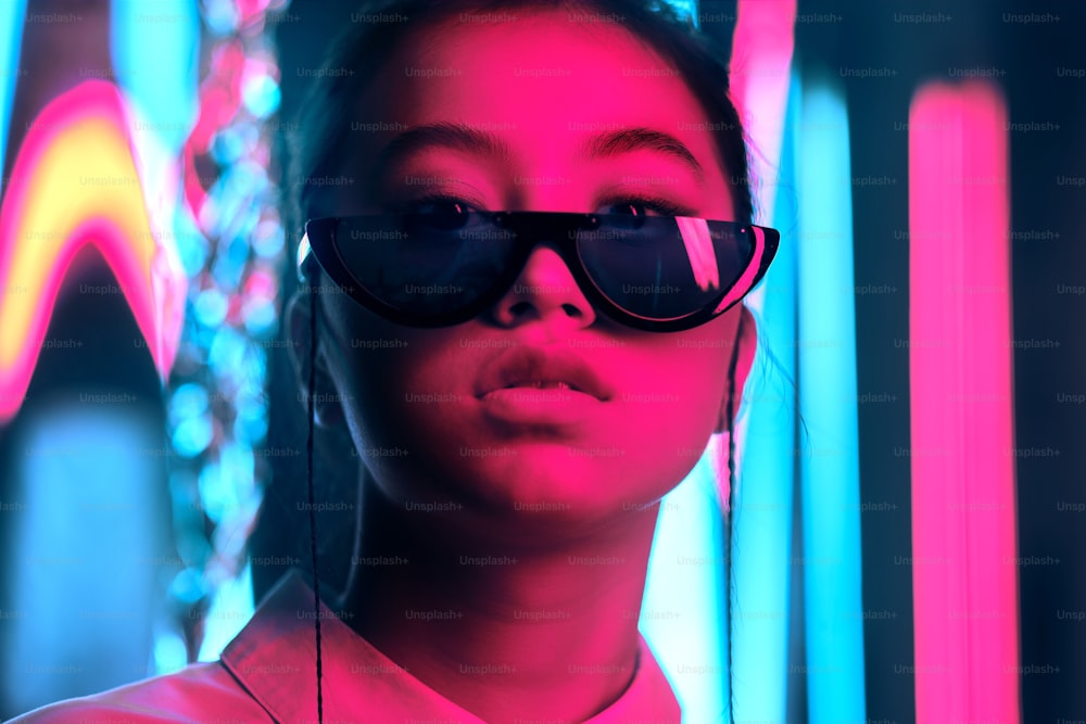スタイリッシュな三日月形のサングラスをかけたアジアの若い10代の女の子のポートレート、赤いアンフブルーのネオンライト。サイバーで未来的なポートレートのコンセプト