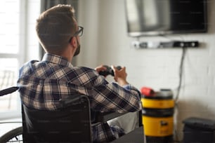 Retrato de la espalda de un joven en silla de ruedas jugando videojuegos en casa. Lleva gafas y camisa a cuadros. Corantino, quédate en casa, concepto de rehabilitación
