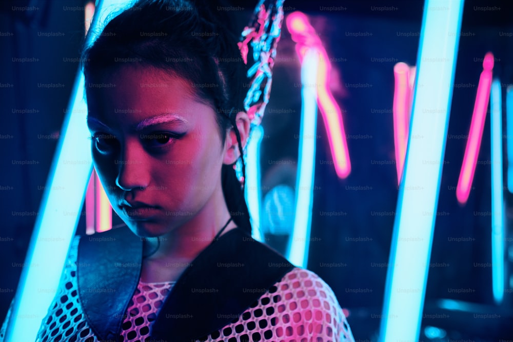 Futuristisches Porträt eines asiatischen Teenagers im Neonlicht mit schwertähnlichen Lampen. Sie ist ein serioses, gewagtes, modisches Cyberpunk-Mädchen, das Netzkleidung und weiße Augenbrauen trägt