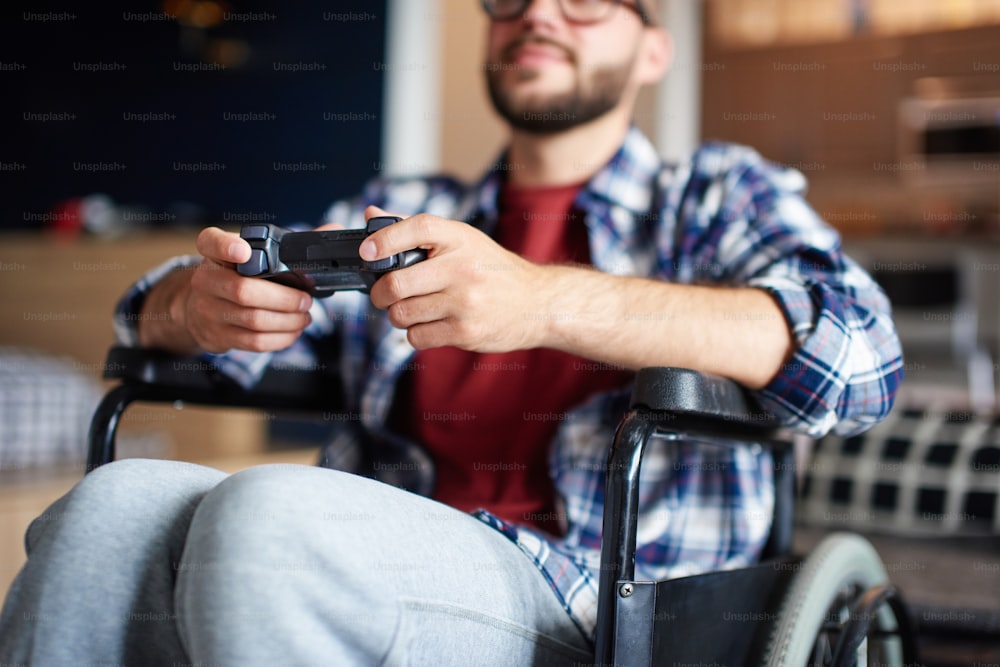Disabled young man playing video games, sitting in wheelchair at home. He is wearing glasses and plaid shirt. Qurantine, stay home, rehabilitation concept