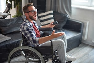 Disabled young man playing video games, sitting in wheelchair at home. He is wearing glasses and plaid shirt. Qurantine, stay home, rehabilitation concept