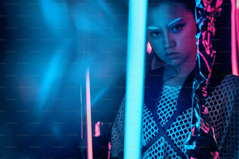 Ritratto futuristico di adolescente asiatico in luce al neon con lampade a forma di spada. Effetti sbavati al neon applicati davanti. È una ragazza alla moda seria, audace, cyberpunk, in abiti netti, sopracciglia bianche
