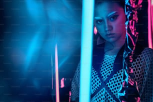 Futuristisches Porträt eines asiatischen Teenagers im Neonlicht mit schwertähnlichen Lampen. Neonverschmierte Effekte auf der Vorderseite. Sie ist ein serioses, gewagtes, modisches Cyberpunk-Mädchen, in Netzkleidung, weißen Augenbrauen