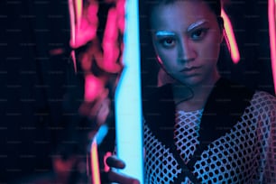 Futuristisches Porträt eines asiatischen Teenagers im Neonlicht, der schwertähnliche Lampen hält. Sie ist ein serioses, gewagtes, modisches Cyberpunk-Mädchen, das Netzkleidung und weiße Augenbrauen trägt