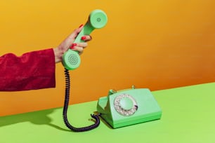オレンジの背景に昔ながらの緑色の電話を持つ女性の手のカラフルな明るい画像、受話器を拾う。ポップアート、ヴィンテージのもの、古いものと現代的なもののコンセプト