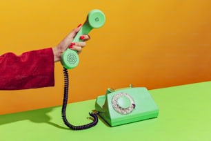 Colorful bright image of female hand holding old-fashioned green colored phone, picking up handset isolated over orange background. Concept of pop art, vintage things, mix old and modernity