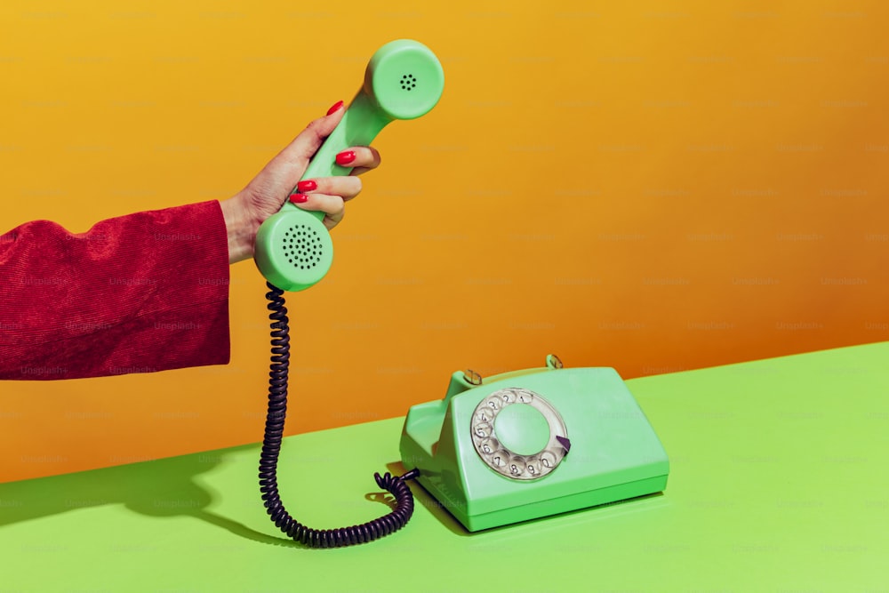 Imagem brilhante colorida da mão feminina segurando o telefone colorido verde à moda antiga, pegando o aparelho isolado sobre o fundo laranja. Conceito de pop art, coisas vintage, mistura de antigo e modernidade