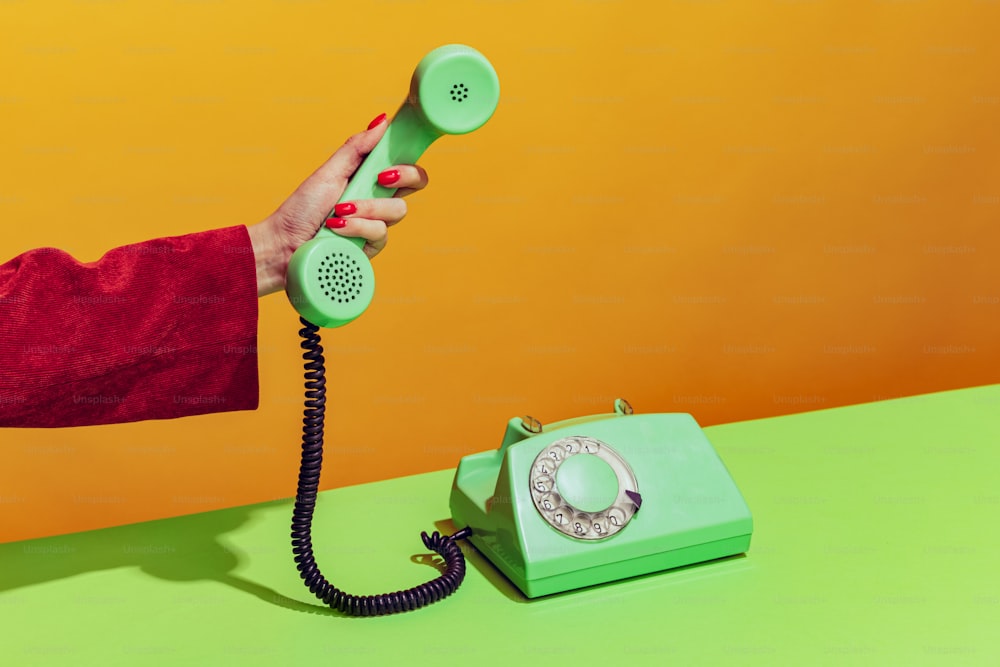 Colorida imagen brillante de la mano femenina sosteniendo un teléfono de color verde anticuado, levantando el auricular aislado sobre un fondo naranja. Concepto de arte pop, cosas vintage, mezcla de lo antiguo y la modernidad