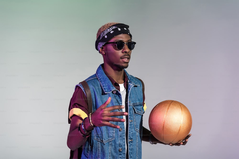 Giovane uomo afroamericano alla moda che tiene la palla dorata da basket isolata sul grigio