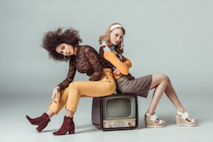 meninas multiculturais estilo retrô sentadas na televisão vintage e olhando para a câmera em cinza