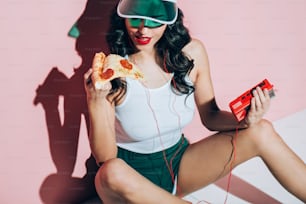 stylish woman in cap with retro music player holding piece of pizza on pink backdrop