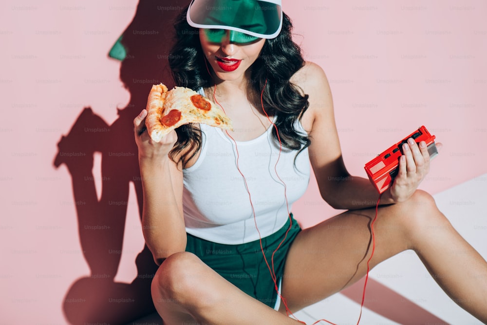 Femme élégante en casquette avec un lecteur de musique rétro tenant un morceau de pizza sur fond rose