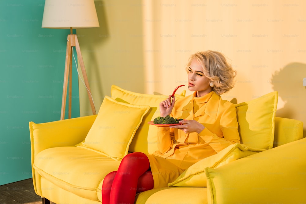 Donna in stile retrò con broccoli sul piatto e peperoncino in mano appoggiata sul divano in appartamento luminoso, concetto di casa delle bambole