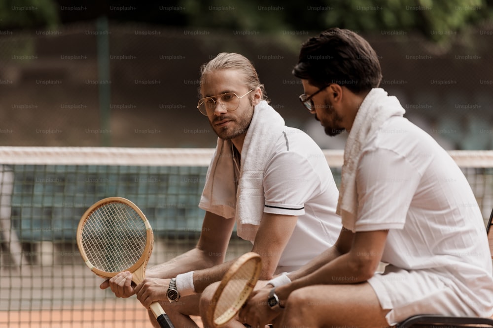 Tennisspieler im Retro-Stil sitzen auf Stühlen mit Handtüchern und Schlägern auf dem Tennisplatz