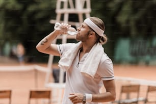 テニスコートで水を飲むハンサムなレトロなスタイルのテニスプレーヤーの側面図