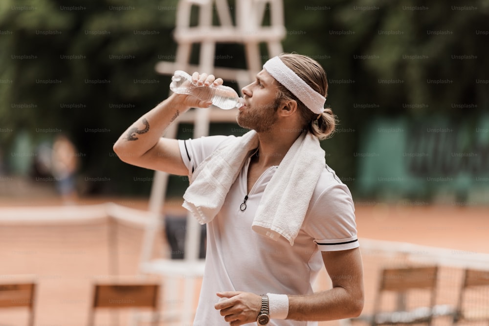 테니스 코트에서 물을 마시는 잘 생긴 복고풍 스타일의 테니스 선수의 측면 모습