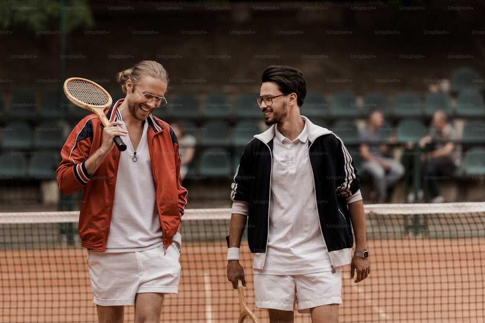 웃고 있는 복고풍 스타일의 테니스 선수들이 테니스 코트에서 라켓을 들고 걷고 있다