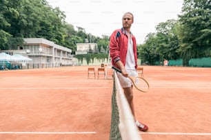 selective focus of handsome tennis player with racket standing at tennis net on court