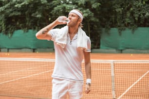 Joueur de tennis fatigué de style rétro avec serviette buvant de l’eau sur le court de tennis