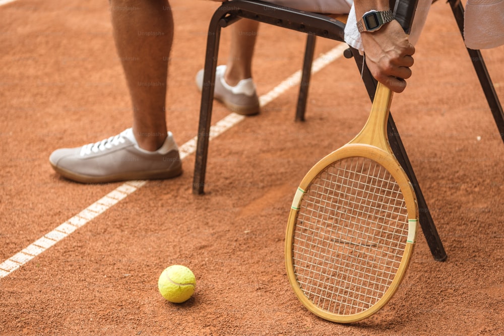 Cropped View Tennisspieler, der sich auf einem Stuhl mit Retro-Holzschläger und Ball auf dem Tennisplatz ausruht