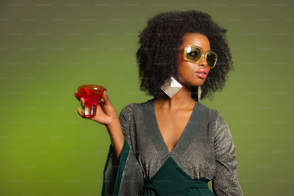 Retro 70s afro fashion woman with green dress and orange cocktail glass. Green wall.