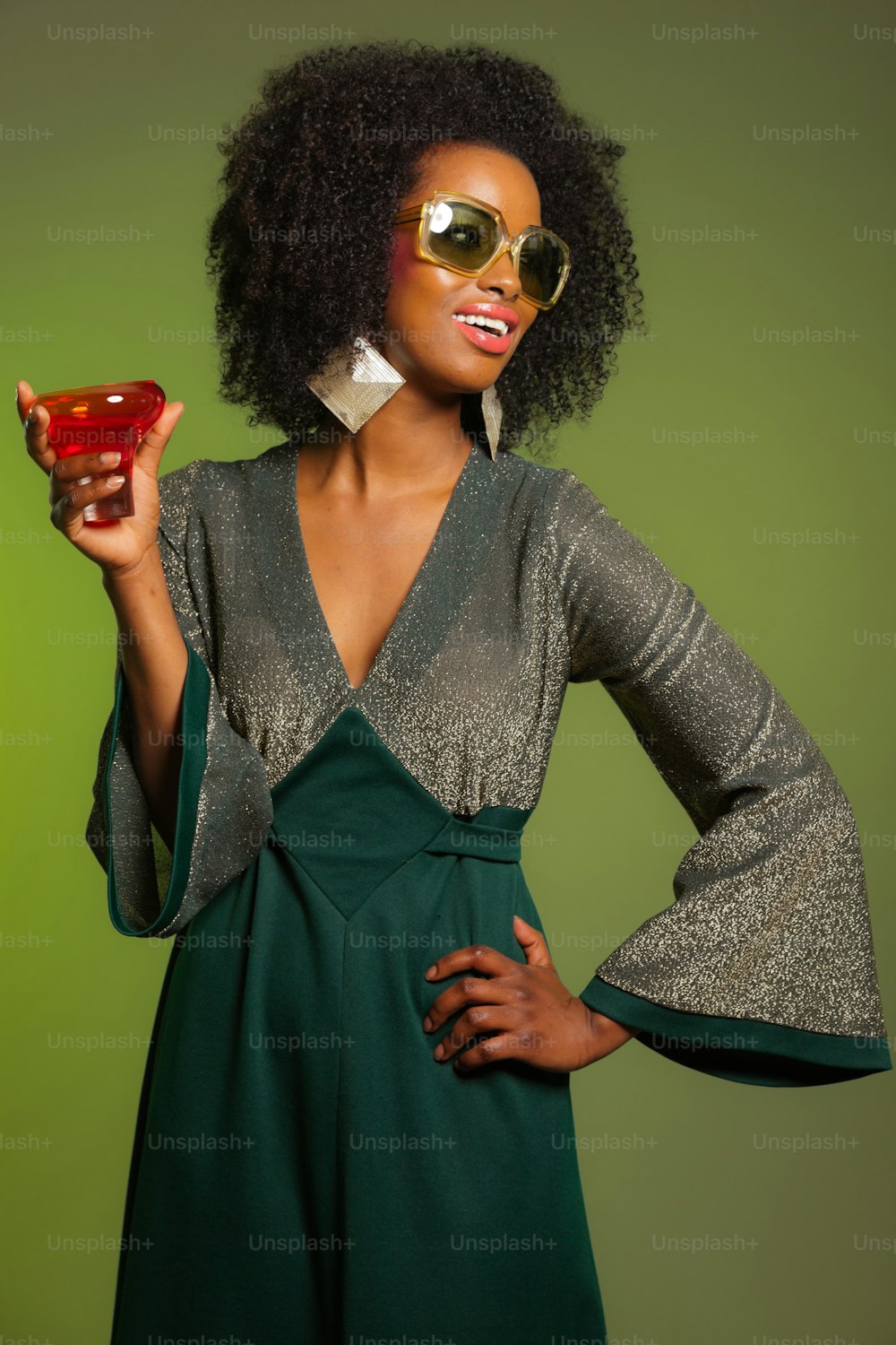 Mulher retrô da moda afro dos anos 70 com vestido verde e copo de coquetel laranja. Parede verde.