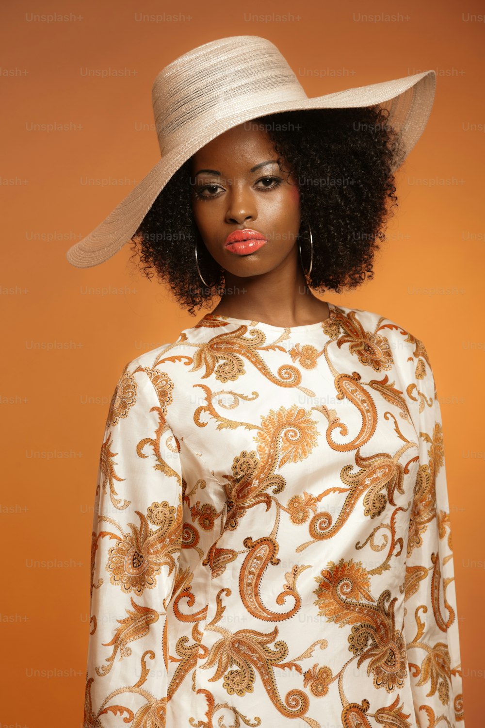 Retro 70s fashion afro woman with paisley dress and white hat. Brown background.