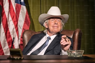 Big boss with white cowboy hat smoking cigar sitting behind desk. American flag in the background.