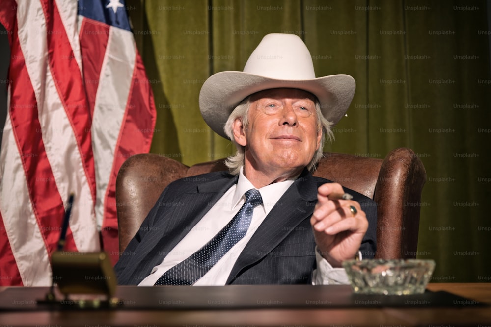 Big boss with white cowboy hat smoking cigar sitting behind desk. American flag in the background.
