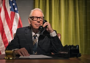 Big boss president wearing glasses calling behind desk with american flag in the background.